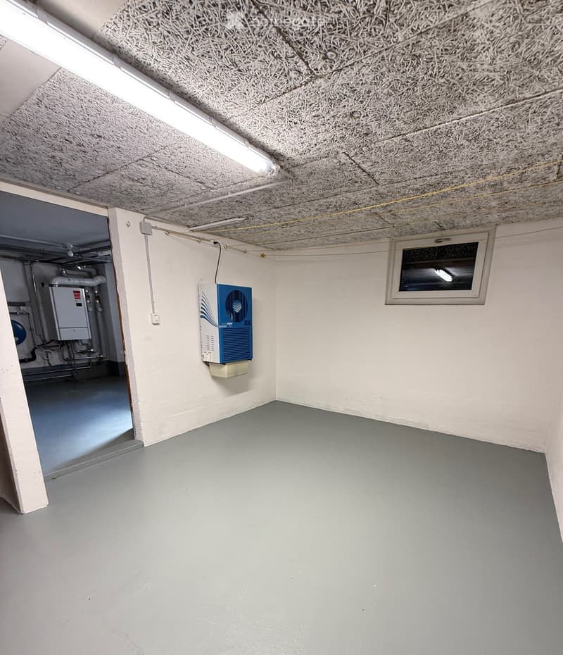 Empty room with grey concrete floor, white walls, and a textured ceiling. Features a small window, a ventilation unit, and an open doorway leading to another area with visible pipes and equipment.