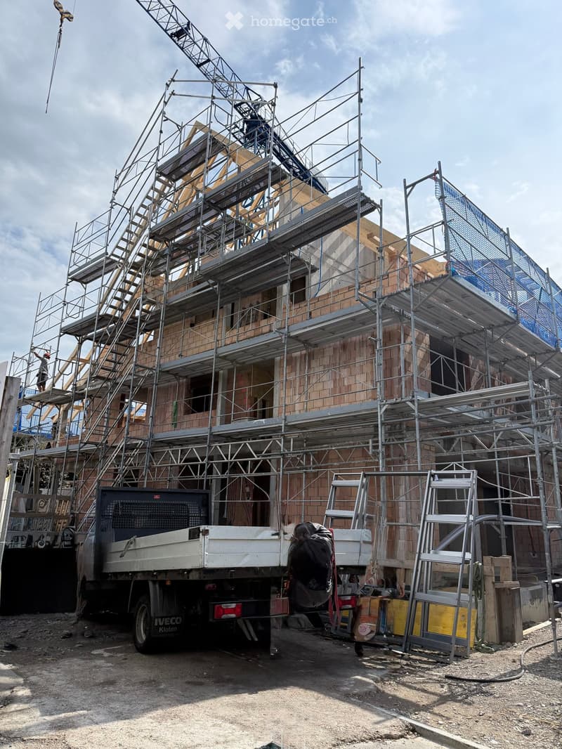 A multi-story building under construction, covered in scaffolding with exposed brickwork and wooden beams. A truck is parked nearby, and construction materials are visible on the ground.
