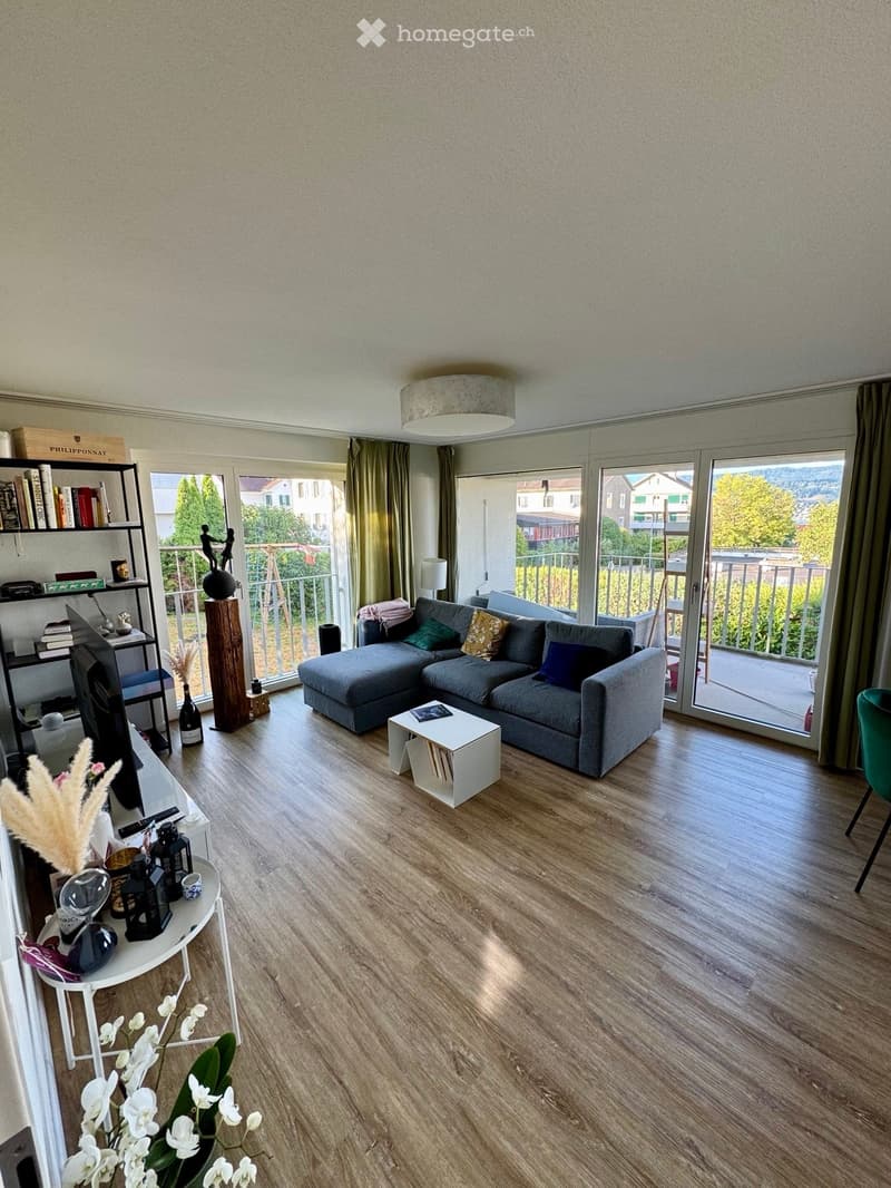 Modern living room with grey sectional sofa, coffee table, bookshelf, and large windows leading to a balcony.