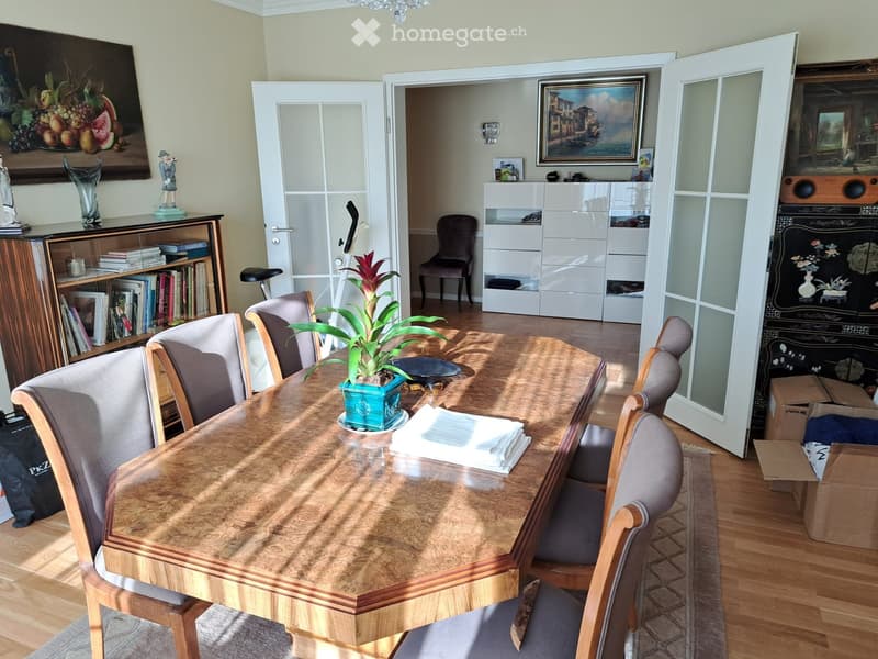 Dining room with a wooden dining table surrounded by six chairs, a potted plant on the table, a bookshelf with books and decorative items, and a white cabinet with drawers.