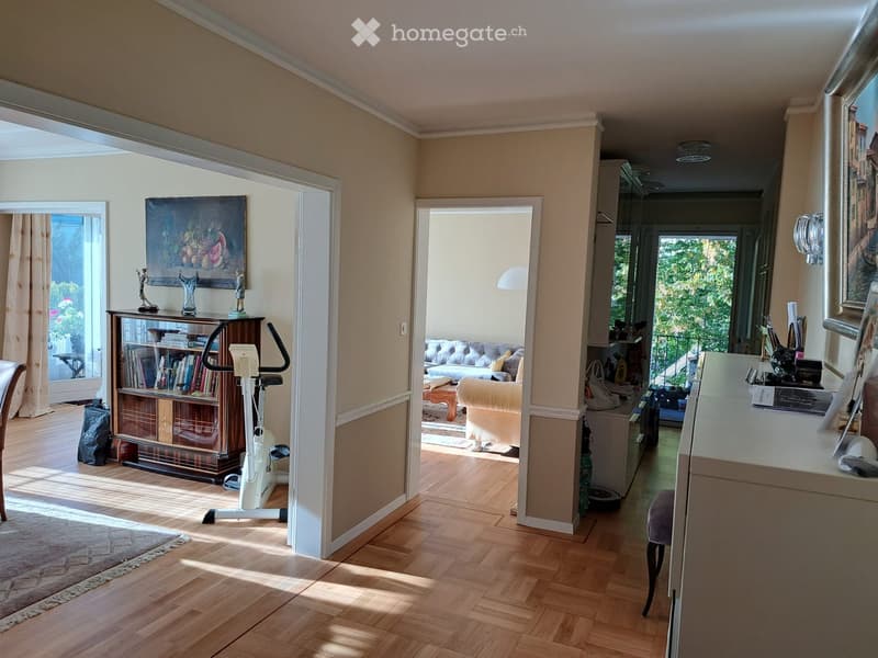 Spacious living area with wooden flooring, large windows, and a view of greenery. Features a sofa, coffee table, exercise bike, bookcase, and a desk with a chair.