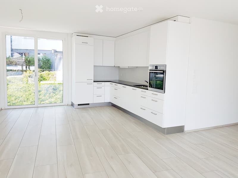 Modern kitchen with white cabinets, light wood flooring, and large windows overlooking a garden.