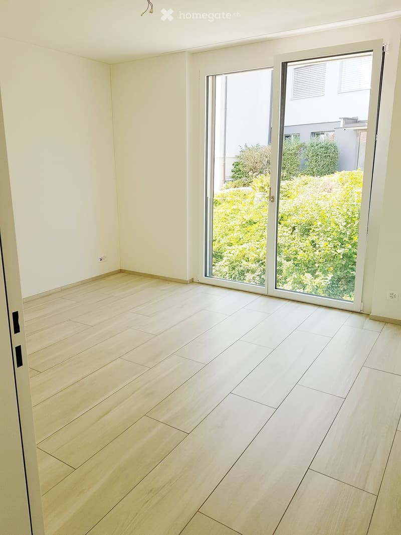 Empty room with light wooden flooring and white walls, featuring large sliding glass doors opening to a garden area.