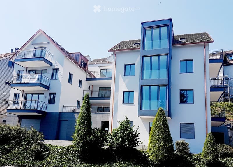 Modern multi-story residential building with white and blue facade, large windows, balconies, and surrounding greenery.
