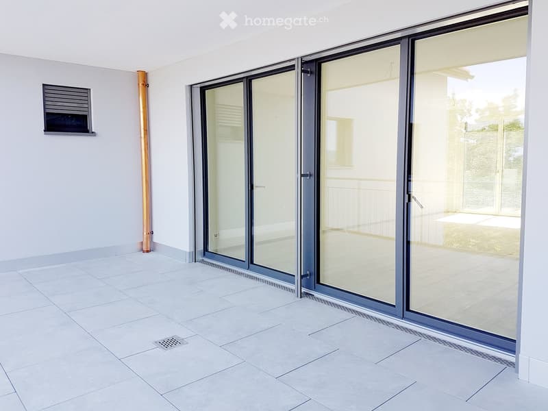 Empty room with tiled floor, large sliding glass doors leading to an outdoor area, and a small window with a shutter on the left wall.