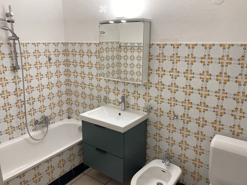 Bathroom with white and yellow floral tiled walls, a bathtub, a sink with a modern faucet, a mirrored cabinet, a bidet, and a toilet.