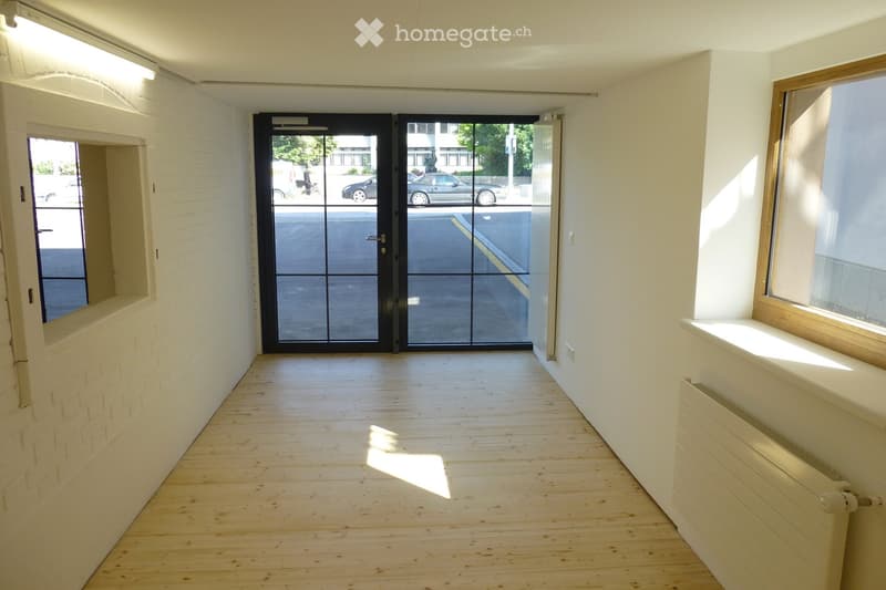 Empty hallway with wooden flooring, white brick wall on one side, large glass door leading outside, and a radiator on the right wall.