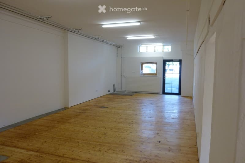 Empty room with wooden flooring, white walls, and a large window at the far end. The space is well-lit with overhead lighting and has a door leading outside.