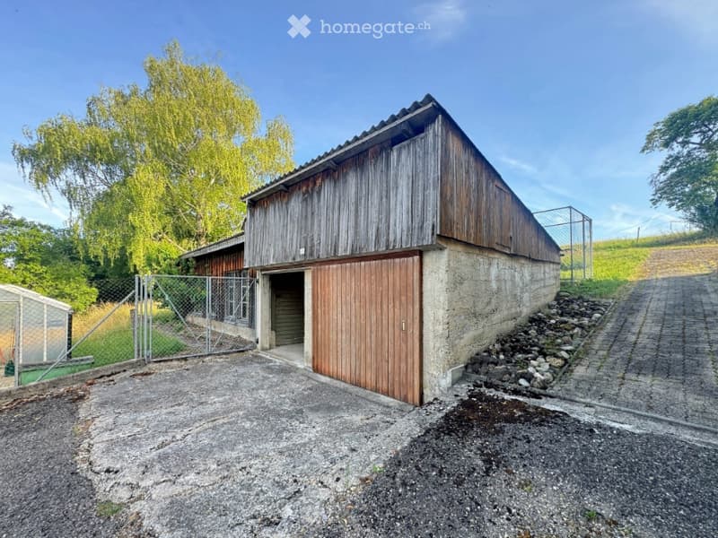 A rustic wooden barn with a concrete base, large wooden doors, and a fenced area in front.