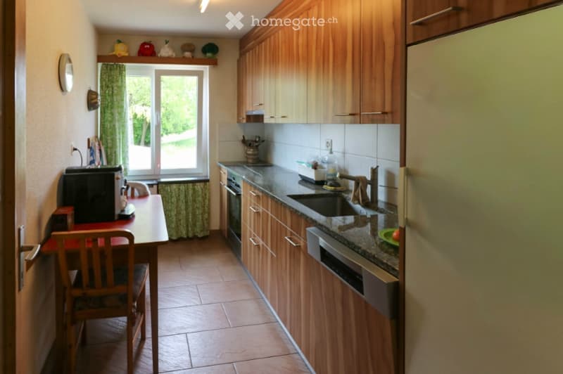 A well-lit kitchen with wooden cabinets, granite countertops, and stainless steel appliances. A small dining area with a table and chair is visible near the window.