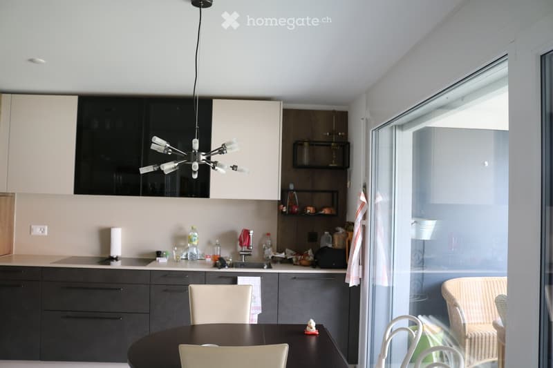 Modern kitchen with light-colored cabinets, dark countertops, and a round dining table with chairs. A hanging light fixture is above the table, and various kitchen items are on the counter.