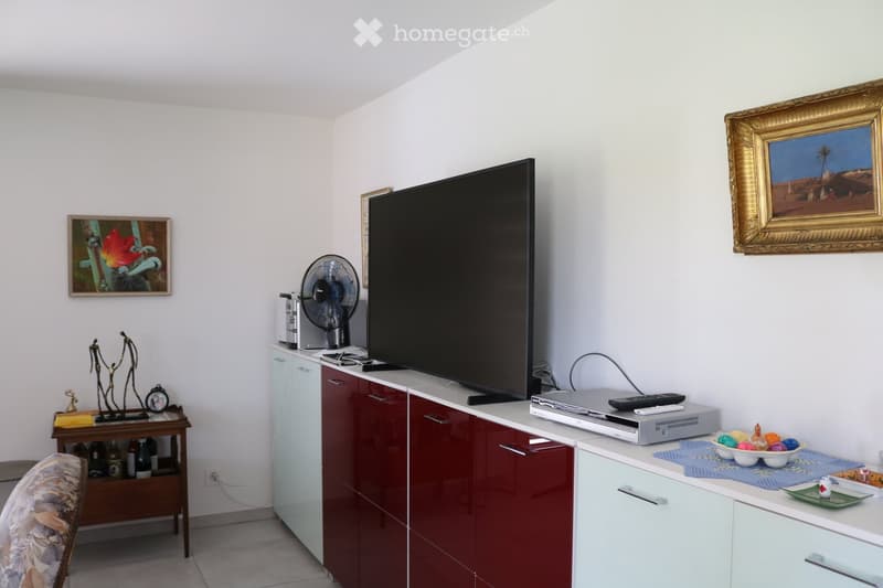 A modern living room with a large flat-screen TV, a small table with decorative items, a fan, and a cabinet with drawers. The room features white walls, a tiled floor, and several framed pictures.