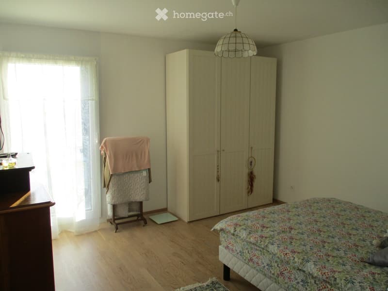 Bright bedroom with wooden flooring, a large bed with floral bedding, a wardrobe, a nightstand with a lamp, and a window with sheer curtains.