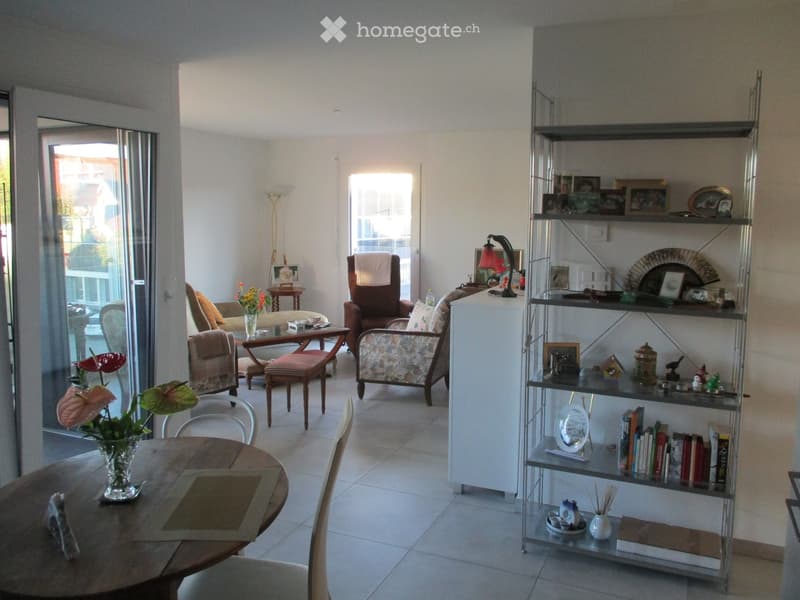 Bright living room with tiled floor, large windows, and sliding glass doors leading to a balcony. Features a round wooden dining table with chairs, a sofa, armchairs, a bookshelf with decorative items, and a lamp.