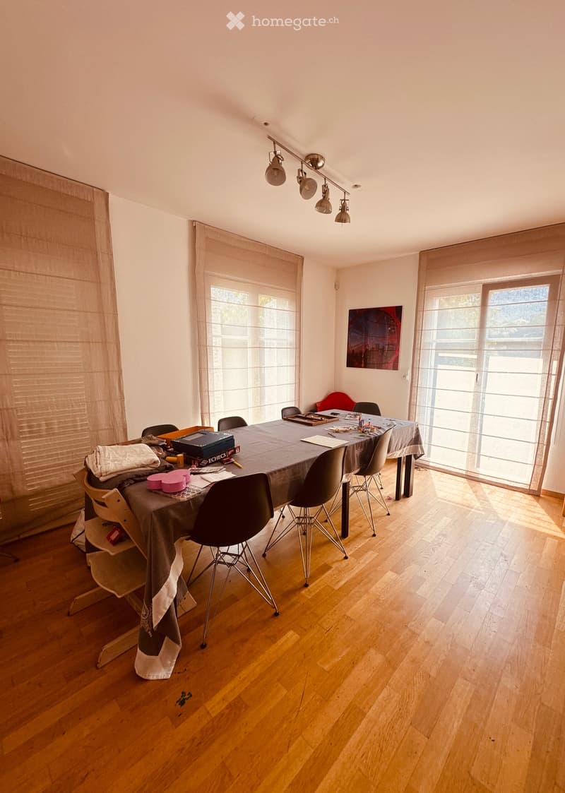 Dining area with wooden floor, large windows with curtains, a table covered with a grey tablecloth, several chairs, and a small stool with folded towels.