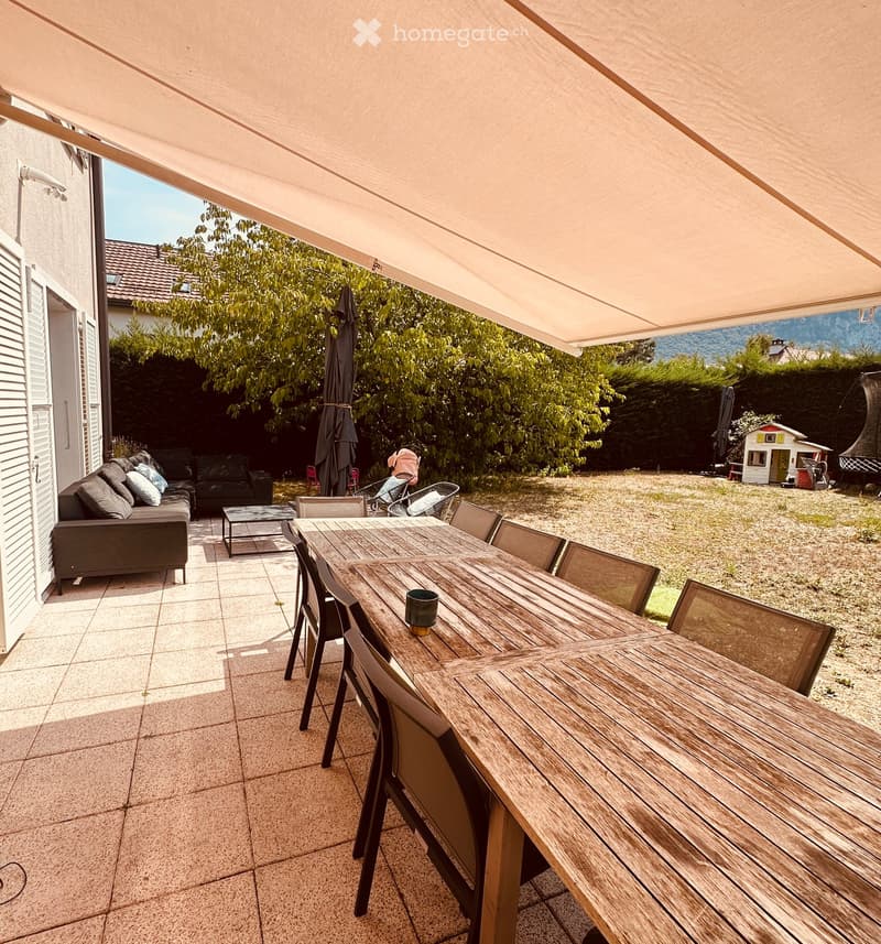 Outdoor patio with a wooden dining table, chairs, a sofa, and a cup on the table. The area is covered by a retractable awning and has a view of a garden with trees and a playhouse.