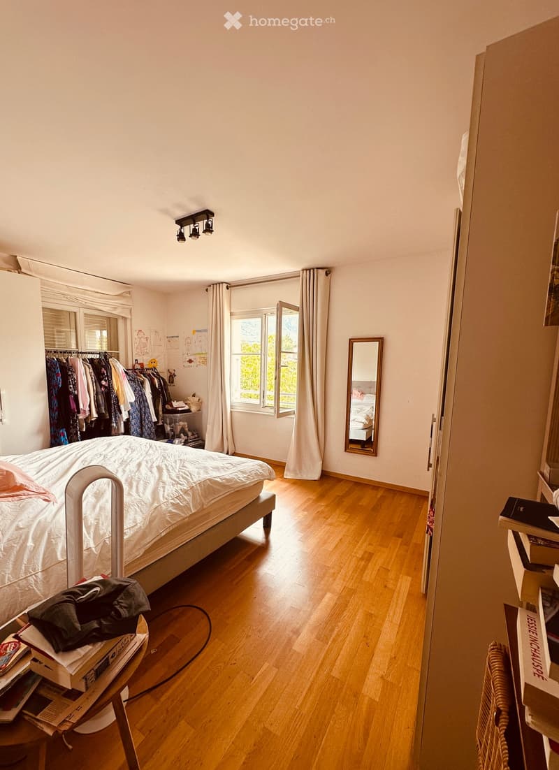 Bright bedroom with wooden flooring, a bed with white bedding, a small table with books and a bag, a mirror, a clothing rack, and large windows with curtains.
