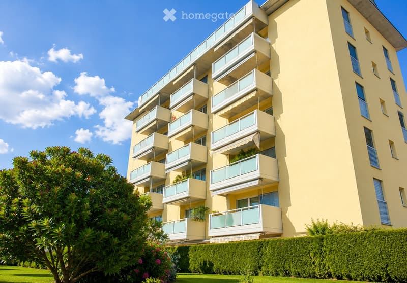 Multi-story residential building with multiple balconies, light yellow facade, and blue shutters. Green lawn and trimmed hedges in front.