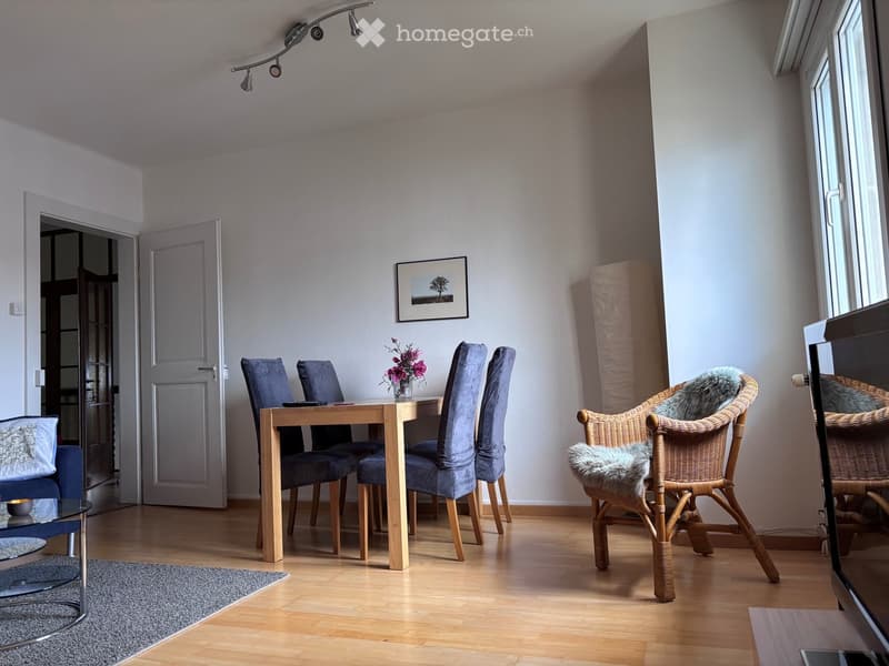 Bright dining area with wooden dining table, four upholstered chairs, wicker chairs with fur cushions, glass coffee table, sofa, and a picture frame on the wall.