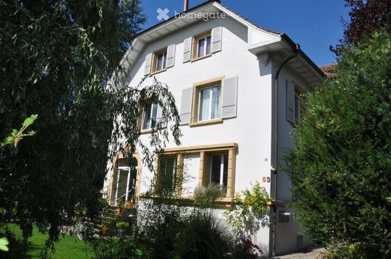 A two-story white house with multiple windows, shutters, and a well-maintained garden.