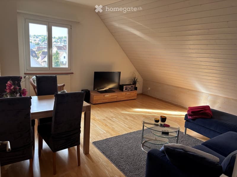 Bright living room with wooden floors, large windows offering a city view, a sofa, a coffee table, a dining table with chairs, and a television on a wooden stand.