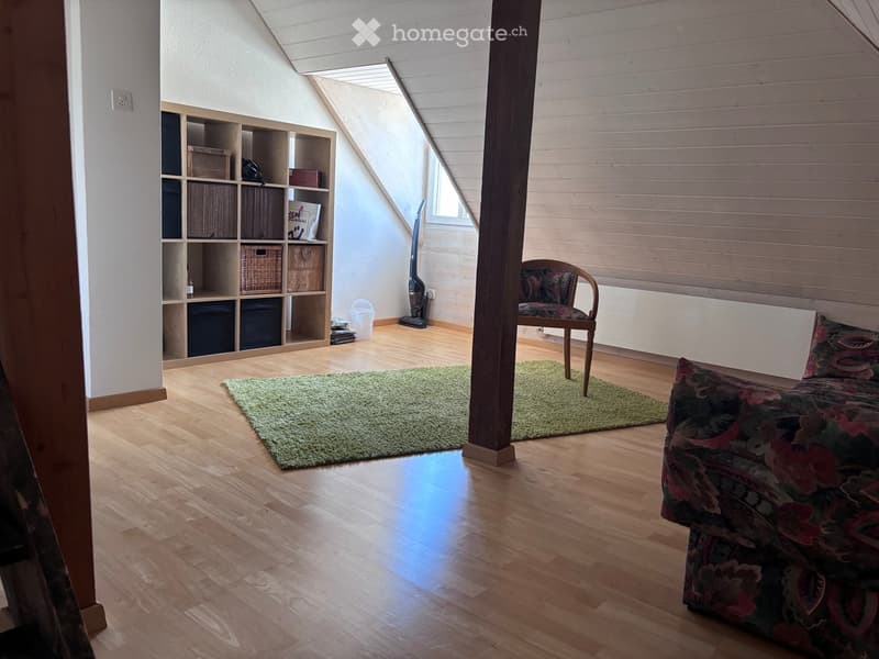 Bright attic room with wooden floors, slanted ceilings, and large windows. Features a patterned sofa, a wooden chair, a green rug, and a wooden shelf with storage boxes.