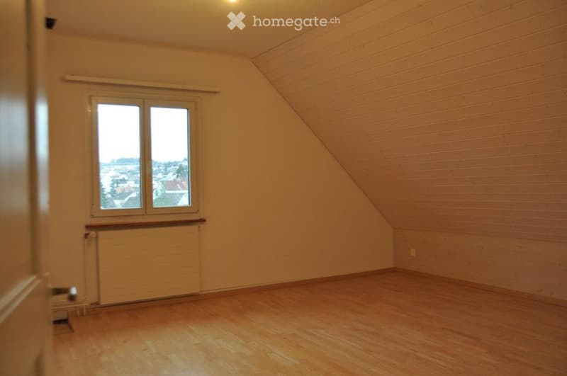 Empty attic room with wooden flooring, slanted wooden ceiling, large window, radiator, and visible light fixture.