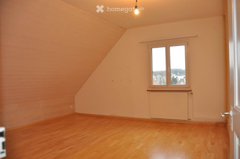 Empty room with wooden flooring, white walls, and a single window with white frame. The ceiling has a sloped design typical of an attic space.