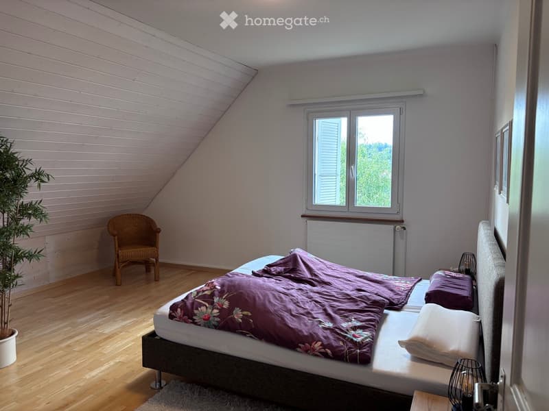 Cozy bedroom with wooden floors, slanted ceiling, large window, bed with purple bedding, wicker chair, small table lamp, and potted plant.
