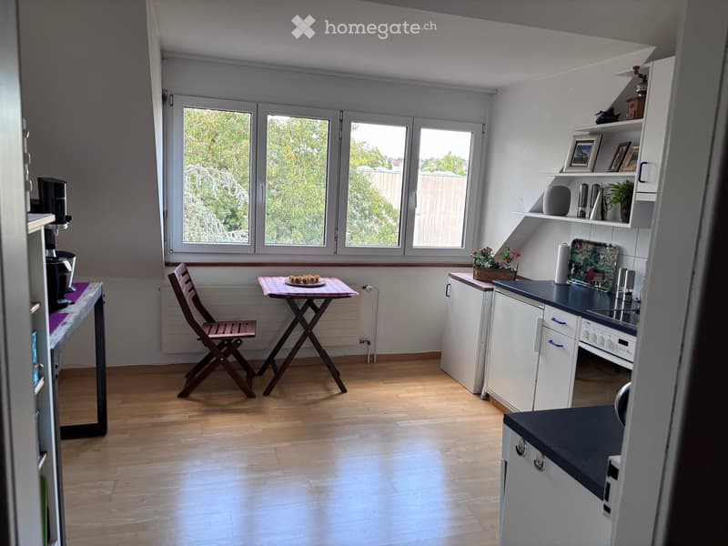 Bright kitchen with wooden flooring, white cabinets, and large windows overlooking greenery. Includes a small table with a chair, coffee maker, and decorative items on shelves.