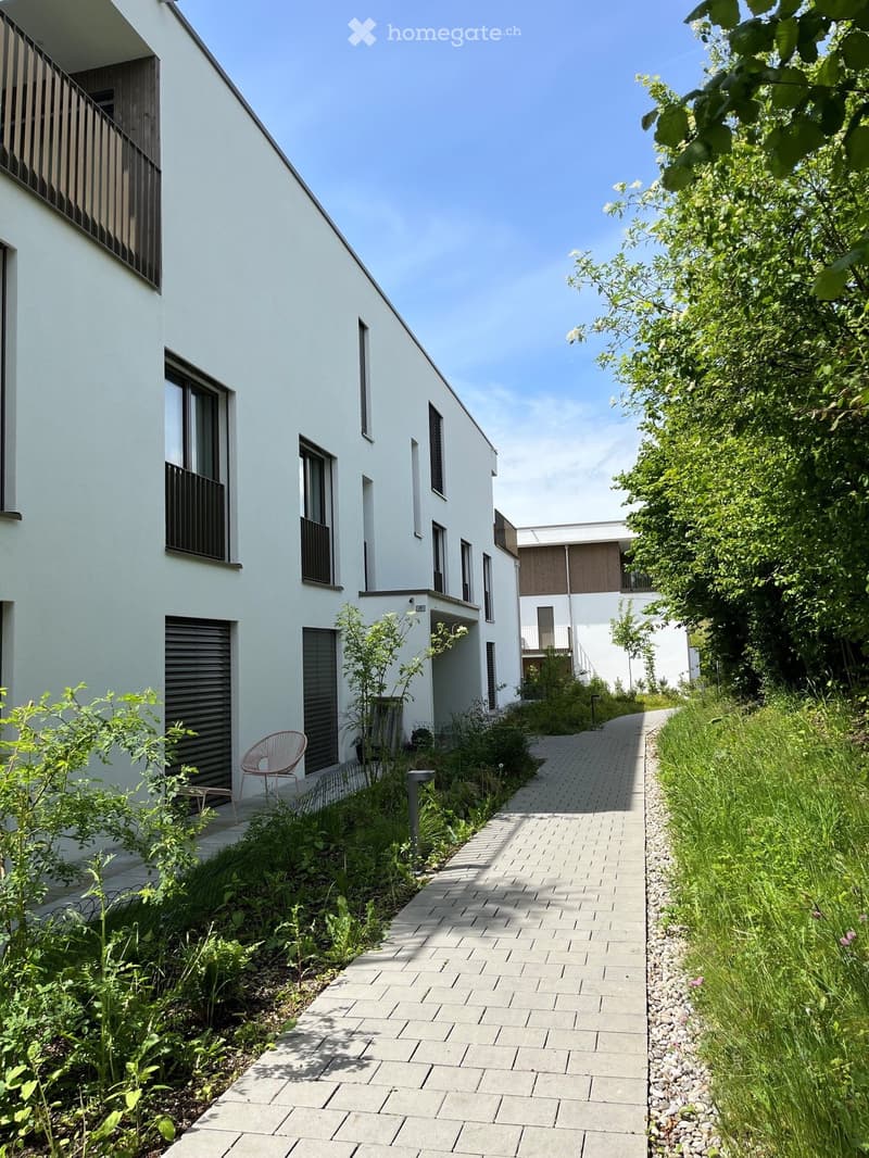 Modern residential complex with a paved walkway, greenery, and contemporary white buildings with large windows and balconies.