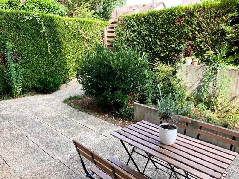 A small, well-maintained garden area with a wooden table, chairs, and potted plants, surrounded by neatly trimmed hedges and a concrete pathway.