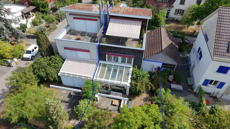 Aerial view of a modern two-story house with a flat roof, large windows, and a spacious garden area. The house features a covered patio and outdoor seating area.