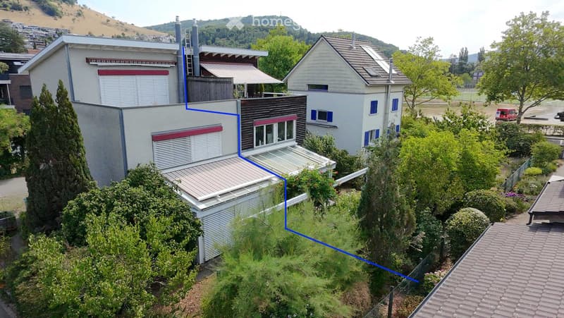 Aerial view of a modern two-story house with white walls, red shutters, and a flat roof extension. The house is surrounded by lush greenery and trees, with neighboring houses visible in the background.