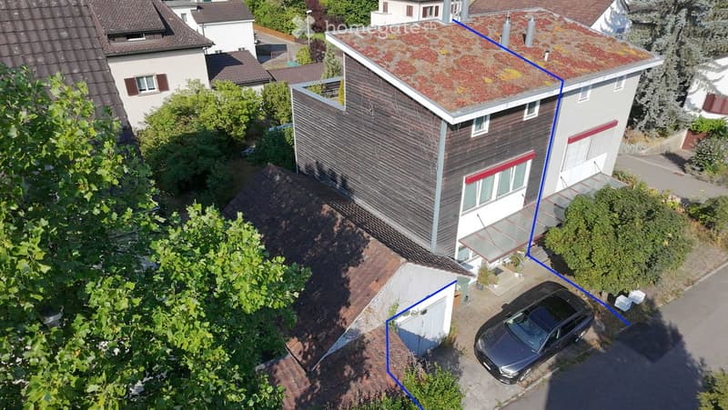 Aerial view of a modern two-story house with a flat roof, surrounded by greenery and neighboring houses. A car is parked in the driveway, and there is a small garage visible.