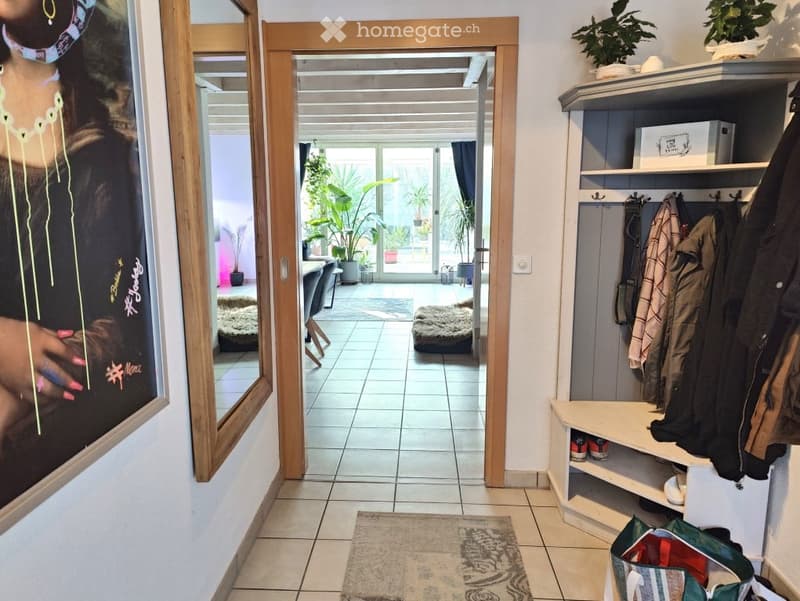 Hallway with tiled floor, coat rack, small rug, and a view into a well-lit living area with plants and furniture.
