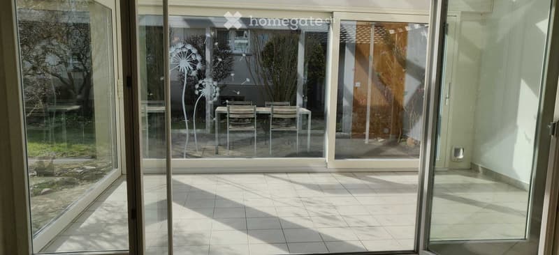 Bright, tiled balcony with large glass doors opening to a garden area, featuring a small table and chairs.