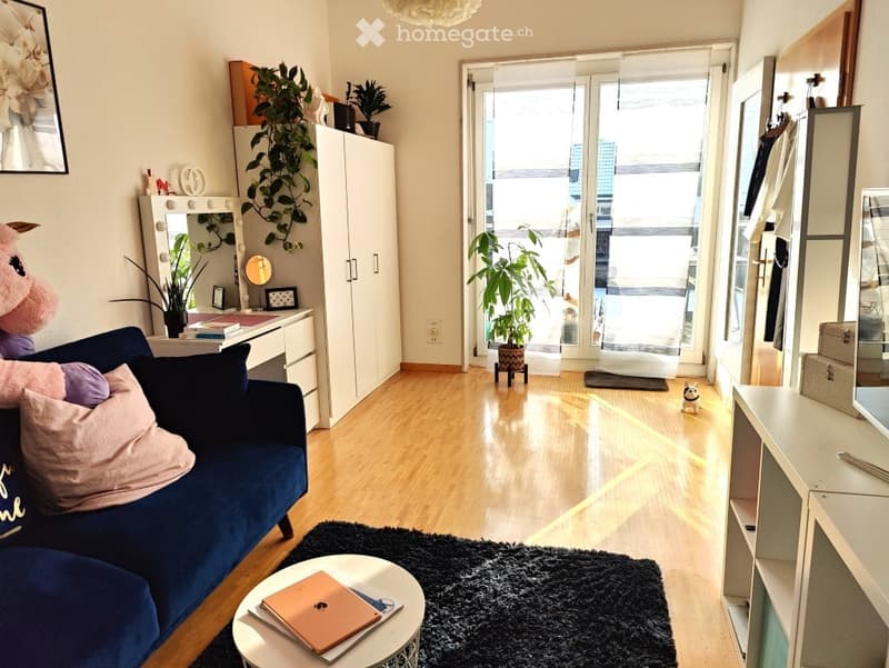 A modern, well-lit living space with a blue sofa, coffee table, laptop, mirror, and plants. The room features a wooden floor, a large window with a view, and a small desk area.
