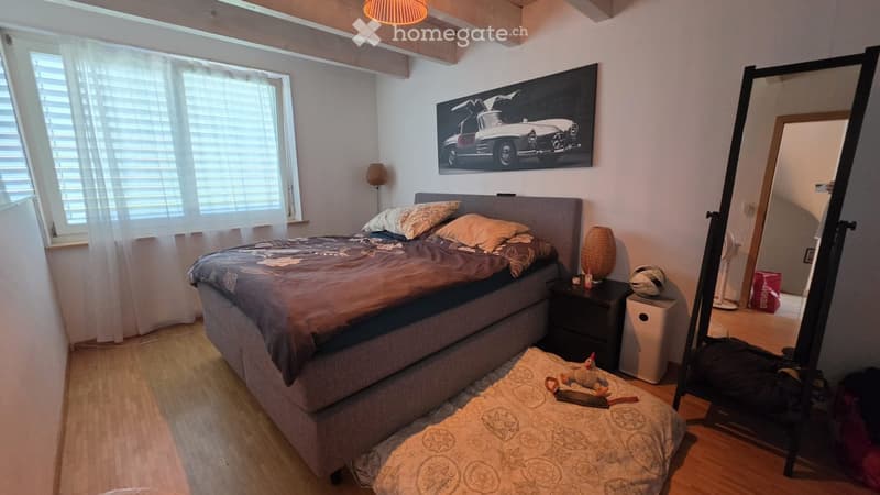 Cozy bedroom with a large bed, nightstand, lamp, mirror, and a large picture on the wall. The room features wooden flooring and large windows with blinds.