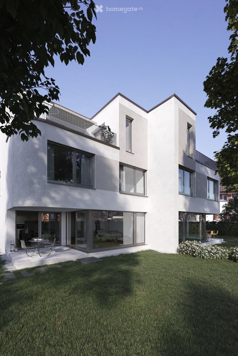 Modern two-story house with white facade, large windows, and a balcony on the upper level. The house features a well-maintained lawn and small patio area with outdoor furniture.
