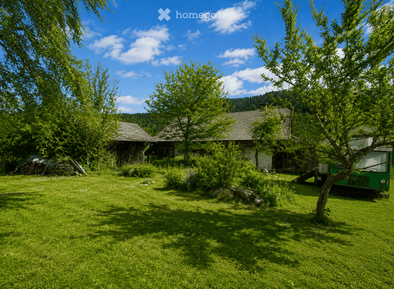 A rural garden area with two wooden houses, lush green grass, trees, and a small green vehicle parked on the right side.