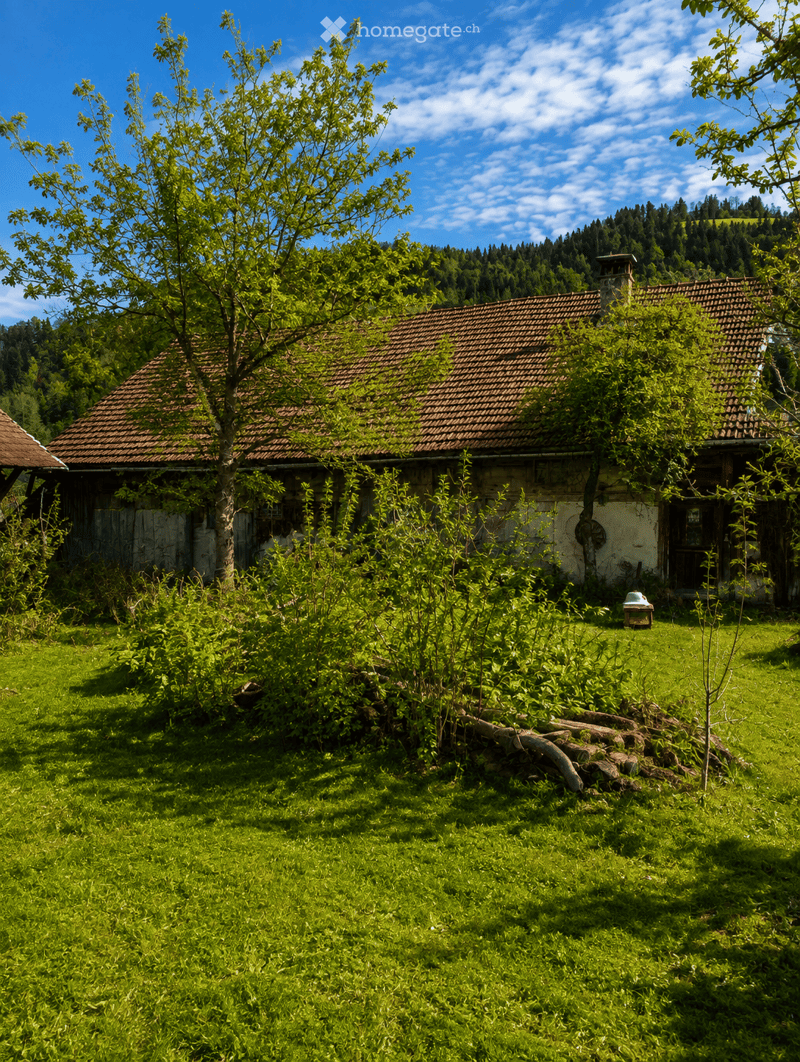 A rustic wooden house with a red-tiled roof surrounded by lush green grass and trees, set in a scenic countryside with hills in the background.