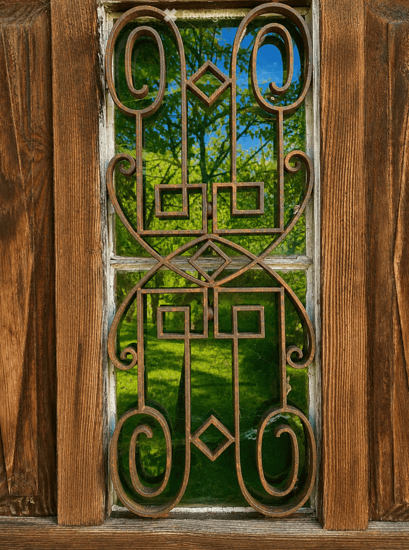 A window with ornate metal bars and glass panes, set in a wooden frame, showing a view of greenery outside.