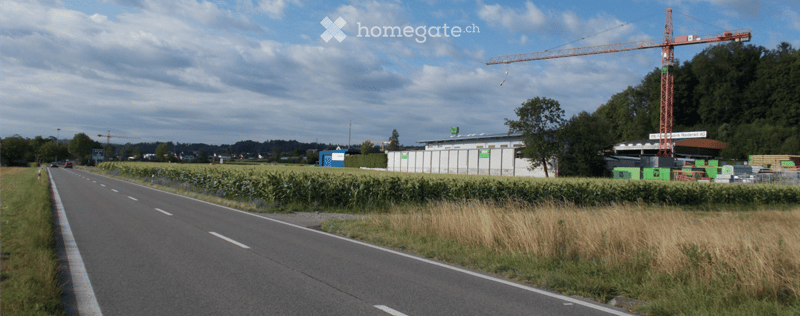Roadside view of a construction site with a large crane, temporary fencing, and a small building with signage.