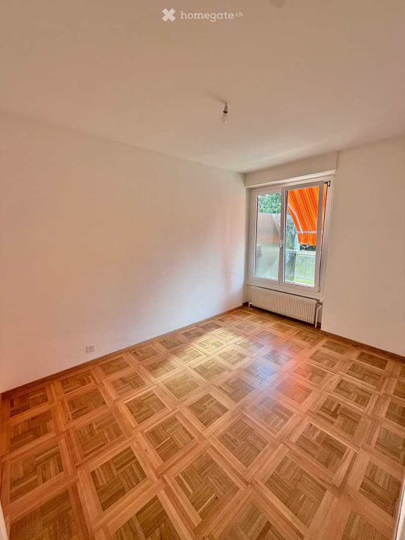 Empty room with wooden parquet flooring, white walls, a radiator, and a large window with a view of the outside.