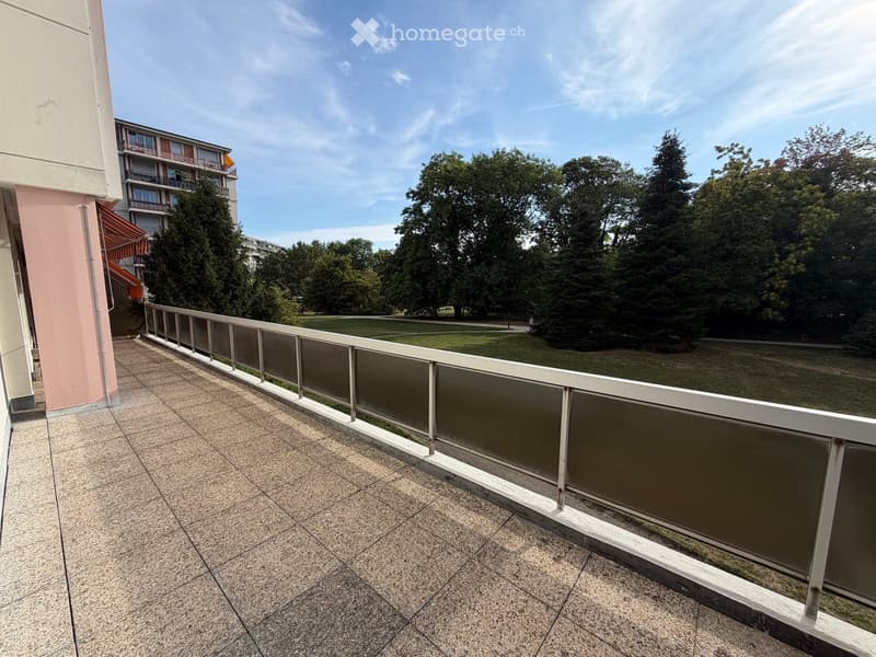 Balcony with tiled floor and white metal railing, overlooking a green park area with trees and a building in the background.