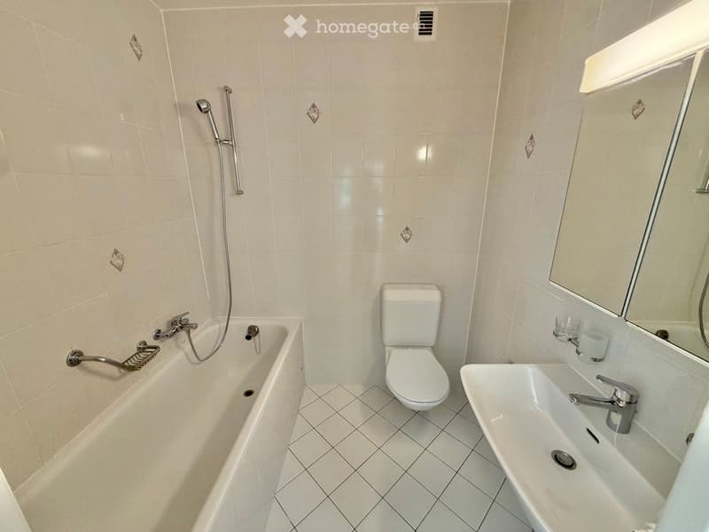 A modern bathroom with a bathtub, toilet, sink, and mirror. The walls and floor are tiled in a light beige color.