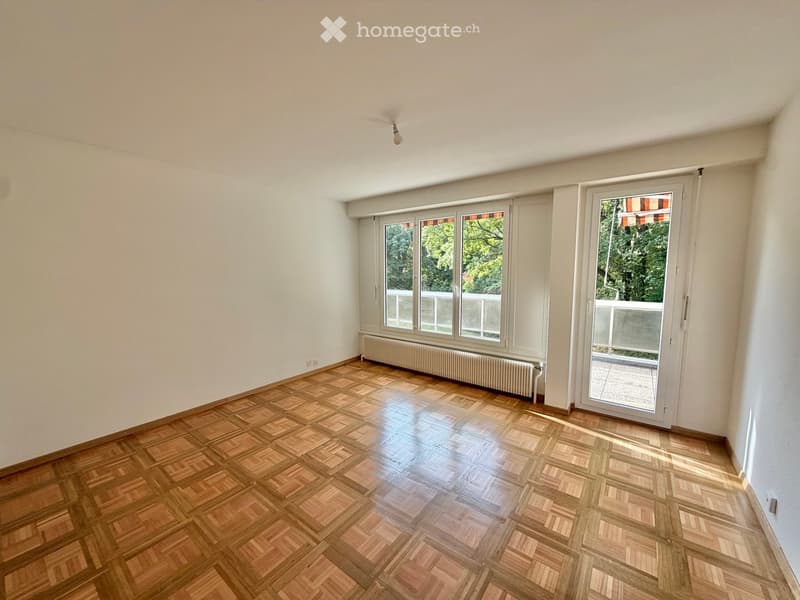 Empty room with wooden parquet flooring, white walls, large windows, and a radiator. No furniture or equipment present.