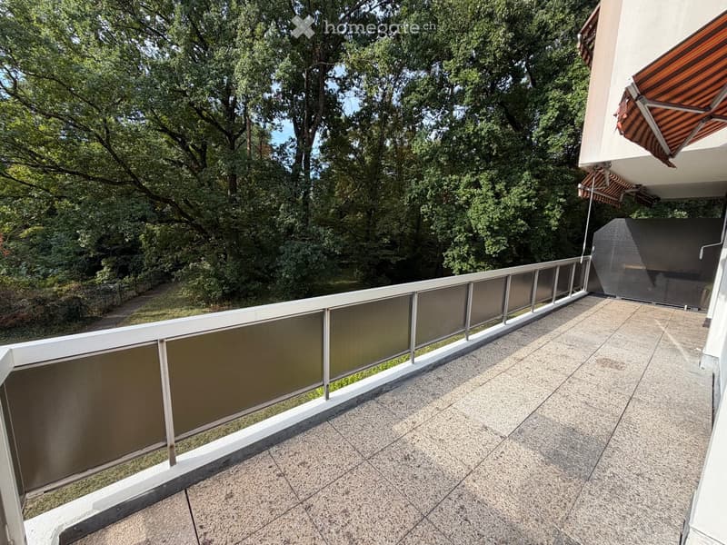 Balcony with tiled floor, glass railing, and a view of a lush green garden area.