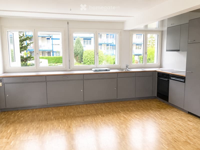 Empty kitchen with grey cabinets, wooden flooring, and large windows overlooking a residential area.